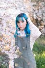 Junge stilvolle Frau mit langen blauen Haaren blickt in die Kamera und trägt einen trendigen Overall und genießt den blühenden Baum, während sie im Frühlingsgarten steht — Stockfoto