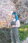 Joven mujer elegante con el pelo largo y azul mirando hacia otro lado usando la fragancia general de moda disfrutando de la floración del árbol mientras está de pie en el jardín de primavera - foto de stock