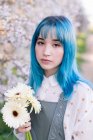 Moderna hembra de moda con cabello azul sosteniendo ramo de flores frescas y mirando a la cámara mientras está de pie en el floreciente jardín de primavera - foto de stock