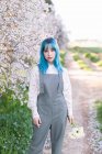 Молодая стильная женщина с длинными голубыми волосами, смотрящая на камеру в модном наряде, наслаждается цветущим деревом, стоя в весеннем саду — стоковое фото
