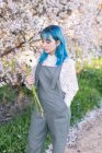 Moderna hembra de moda con cabello azul sosteniendo y mirando el ramo de flores frescas mientras está de pie en el floreciente jardín de primavera - foto de stock