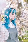 Молода стильна жінка з довгим блакитним волоссям з закритими очима в модному загальному, насолоджуючись ароматом квітучого дерева, стоячи в весняному саду — стокове фото