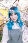 Молода стильна жінка з довгим синім волоссям дивиться на камеру в модному загальному, насолоджуючись квітучим деревом, стоячи в весняному саду — стокове фото