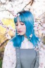 Молодая стильная женщина с длинными голубыми волосами с закрытыми глазами, одетая в модный общий аромат цветущего дерева, стоя в весеннем саду — стоковое фото