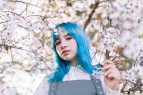 Giovane femmina elegante con lunghi capelli blu guardando la fotocamera indossa trendy generale godendo albero in fiore mentre in piedi in giardino primaverile — Foto stock