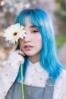 Moderna fêmea moderna com cabelo azul segurando um olho de cobertura de flor fresca e olhando para a câmera enquanto está em pé no jardim de primavera florescendo — Fotografia de Stock