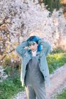Joven mujer elegante tocando el pelo largo y azul mirando a la cámara usando moda en general disfrutando del árbol floreciente mientras está de pie en el jardín de primavera - foto de stock
