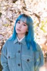 Молода стильна жінка з довгим синім волоссям з руками в кишені дивиться на камеру в модному загальному, насолоджуючись квітучим деревом, стоячи в весняному саду — стокове фото