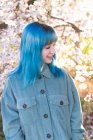 Junge stylische Frau mit langen blauen Haaren mit den Händen in der Tasche, die wegschauen und trendigen Overall tragen, der blühenden Baum genießt, während sie im Frühlingsgarten steht — Stockfoto
