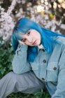 Triste modèle féminin millénaire avec des cheveux bleus dans une tenue élégante en regardant la caméra soigneusement tout en étant assis sur l'herbe verte près de l'arbre en fleurs dans le jardin de printemps — Photo de stock