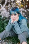 Грустная модель тысячелетия с голубыми волосами в стильном наряде, задумчиво смотрящая на камеру, сидя на зеленой траве возле цветущего дерева в весеннем саду — стоковое фото
