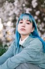Sad millennial female model with blue hair in stylish outfit looking at camera thoughtfully while sitting on green grass near blooming tree in spring garden — Stock Photo