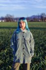 Продумана молода жінка з блакитним волоссям дивиться на камеру, одягнену в модну куртку, що стоїть в зеленому полі в сонячний вечір — стокове фото