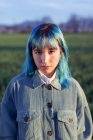 Jeune femme réfléchie aux cheveux bleus regardant la caméra vêtue d'une veste tendance debout dans un champ vert dans une soirée ensoleillée — Photo de stock