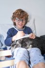 Menino loiro com óculos escrevendo em um caderno sentado na cama com seu cachorro — Fotografia de Stock
