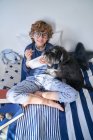 Blond boy with glasses writing in a notebook sitting on the bed with his dog — Stock Photo