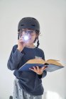 Criança loira em pijama com um capacete uma lanterna e um livro jogando pesquisa — Fotografia de Stock