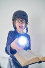 Blondes Kind im Schlafanzug mit Helm, Taschenlampe und Buch beim Recherchieren — Stockfoto