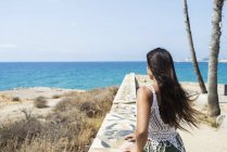 Visão traseira de uma mulher hispânica de cabelos longos apoiada em uma parede na praia enquanto olha para longe para o horizonte — Fotografia de Stock
