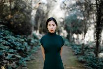 Молода азіатка в модному одязі в зелених кущах і дивиться на камеру в літньому саду — стокове фото