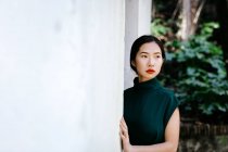 Joven mujer asiática en vestido de moda en arbustos verdes y mirando hacia otro lado apoyado en la pared blanca en el jardín envejecido - foto de stock