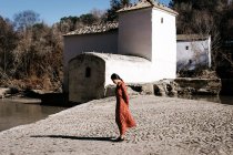Vista lateral de mujer asiática en vestido elegante de pie en camino de piedra áspera cerca de lago tranquilo y edificio antiguo en el campo - foto de stock