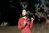 Hermosa mujer asiática con los ojos cerrados y sombra de ramita de plantas en la cara de pie contra el lago en el campo - foto de stock