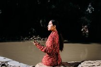 Vista lateral de la mujer asiática con estilo mirando hacia otro lado mientras está sentado en una valla de piedra que sostiene la rama delgada cerca del lago tranquilo - foto de stock