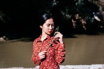 Elegante mulher asiática com olhos fechados segurando ramo fino em pé perto do lago pacífico no dia sem nuvens no campo — Fotografia de Stock
