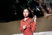Stylish asian woman with closed eyes holding thin branch standing near peaceful lake on cloudless day in countryside — Stock Photo