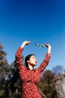 Stylish asian woman with closed eyes holding thin branch in raised arms while standing near peaceful lake on cloudless day in countryside — Stock Photo