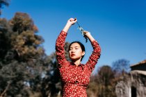 Elegante mulher asiática com olhos fechados segurando ramo fino em braços levantados enquanto estava perto do lago pacífico no dia sem nuvens no campo — Fotografia de Stock