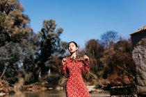 Stylish asian woman with closed eyes holding thin branch while standing near peaceful lake on cloudless day in countryside — Stock Photo