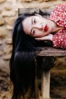 Asiatin in trendiger floraler Kleidung liegt auf alter Holzbank und blickt in die Kamera gegen Steinmauer — Stockfoto