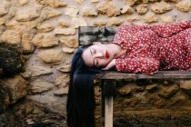 Donna asiatica in abiti floreali alla moda sdraiata su una vecchia panchina di legno con gli occhi chiusi contro il muro di pietra — Foto stock