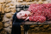 Asian woman in trendy floral clothes lying on old lumber bench and looking at camera against stone wall — Stock Photo