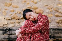 Vista laterale di attraente donna asiatica in abito floreale abbracciando le ginocchia e guardando la fotocamera mentre seduto su una panchina di legno squallido contro muro di pietra — Foto stock