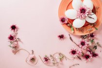 Зверху видніються витончені квіти, розміщені біля тарілки з яйцями курки на рожевому фоні. — стокове фото