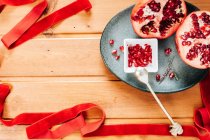 Top view of red ribbon placed on lumber table near plate with halves of ripe pomegranate and spoon — Stock Photo