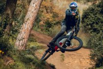 Unrecognizable man in helmet, gloves and protection glasses jumping doing whip trick downhill during mountain biking practice in wood forest — Stock Photo
