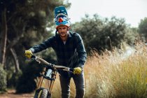 Bonito homem de capacete andando olhando para a câmera enquanto carrega bicicleta de montanha após a prática no prado — Fotografia de Stock