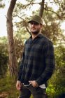 Portrait de bel homme brunet en chemise à carreaux et casquette de baseball debout dans la nature en arrière-plan regardant la caméra — Photo de stock