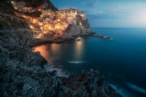 Increíble paisaje con pequeña ciudad con luces de colores en la costa rocosa lavado por el agua del océano tranquilo por la noche - foto de stock