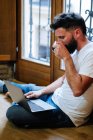 Homem barbudo bebendo bebida quente e lendo dados do laptop enquanto sentado no chão e trabalhando em projeto remoto em casa — Fotografia de Stock