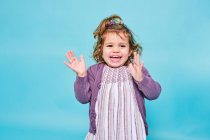 Petit enfant joyeux en robe violette et cardigan tricoté souriant à la caméra et applaudissant tout en restant seul sur fond bleu clair dans un studio moderne — Photo de stock