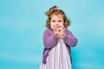 Cheerful small child in purple dress and knitted cardigan smiling at camera and clapping while standing alone against light blue background in modern studio — Stock Photo