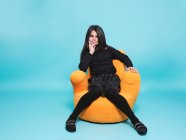 Hübsches Mädchen in schwarzer Freizeitkleidung, die Hand am Kinn liegend, in die Kamera blickend, während es sich in leuchtend gelben Sesseln vor hellblauem Hintergrund in einem modernen Studio entspannt — Stockfoto