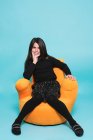 Hübsches Mädchen in schwarzer Freizeitkleidung, die Hand auf dem Kinn liegend lächelnd in die Kamera blickend, während es sich in leuchtend gelbem Sessel vor hellblauem Hintergrund in einem modernen Studio entspannt — Stockfoto