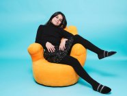 Low angle of happy preteen girl in black casual clothes smiling looking at camera while relaxing in bright yellow armchair hand shape against light blue background in contemporary studio — Stock Photo