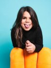 Menina pré-adolescente feliz em roupas casuais pretas sorrindo furando a língua para fora para a câmera enquanto se inclina atrás de poltrona amarela contra fundo azul claro no estúdio contemporâneo — Fotografia de Stock